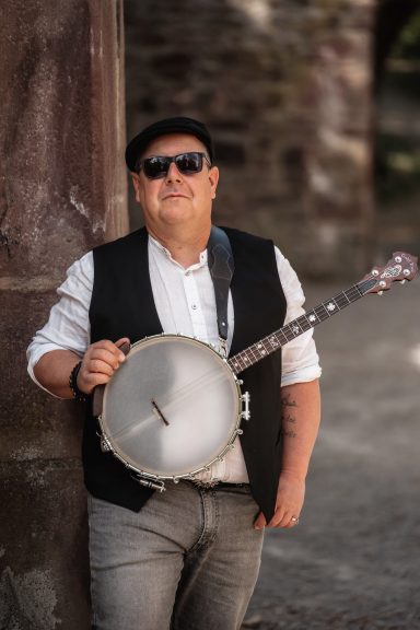 Mann in Weste und Hut hält eine Banjo, lächelt und steht an einer Mauer.