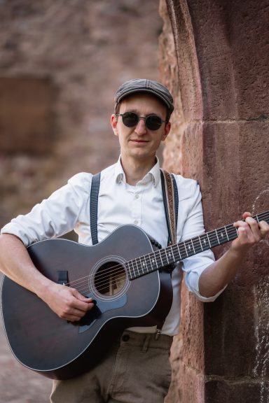 Mann in Sonnenbrille und Mütze spielt Gitarre vor einer Steinmauer.