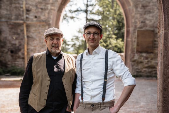 Zwei Männer in historischen Kleidern stehen vor einer alten Mauer.