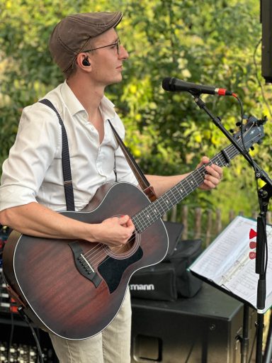 Mann mit Gitarre und Mikrofon, trägt Hemd und Hut, spielt im Freien.