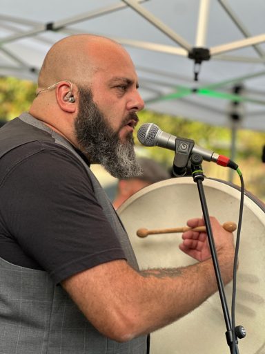 Mann mit Bart, der auf einer Bühne singt und ein Trommelinstrument spielt.