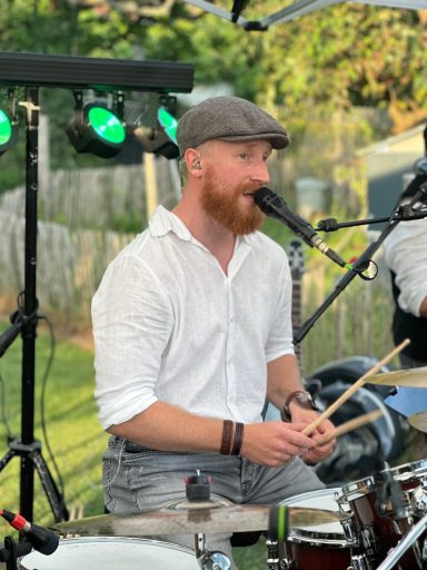 Ein Musiker mit rotem Bart und Mütze spielt Schlagzeug und singt im Freien.