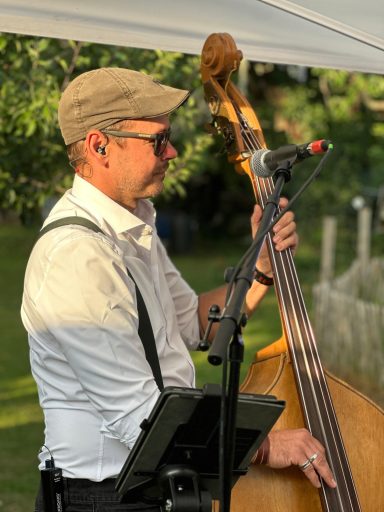 Mann spielt Kontrabass, trägt eine Mütze und eine weiße Hemd mit Hosenträgern.